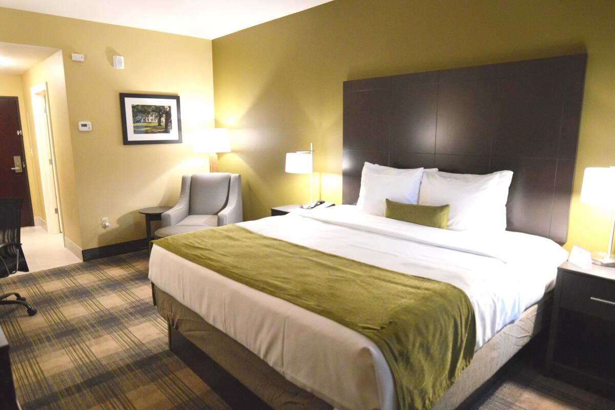 A hotel room with a large bed, a chair, a desk, and a nightstand with a lamp.