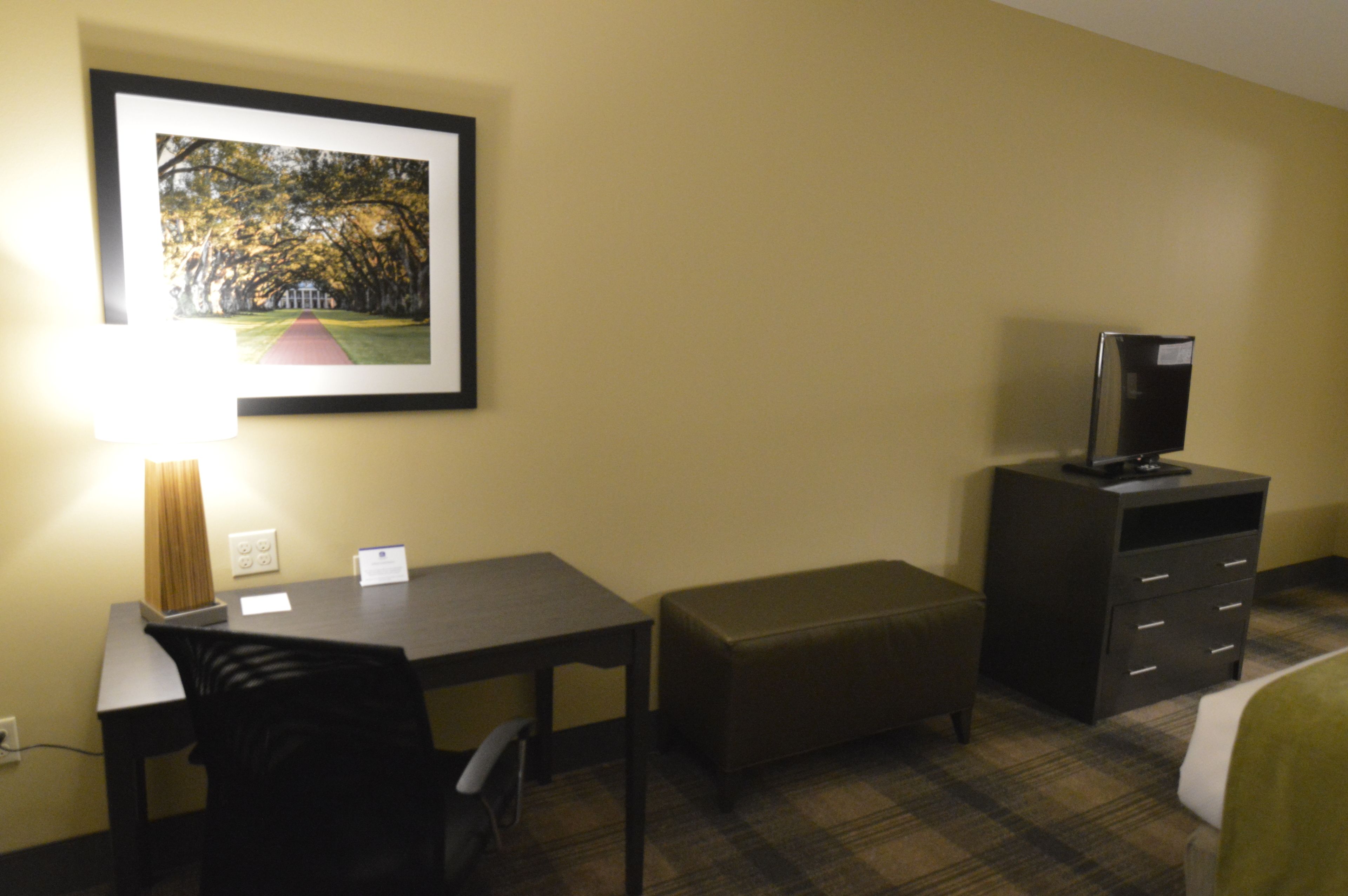 Quarto de hotel com escrivaninha, uma cadeira, um banco, uma TV sobre um armário e um quadro na parede.