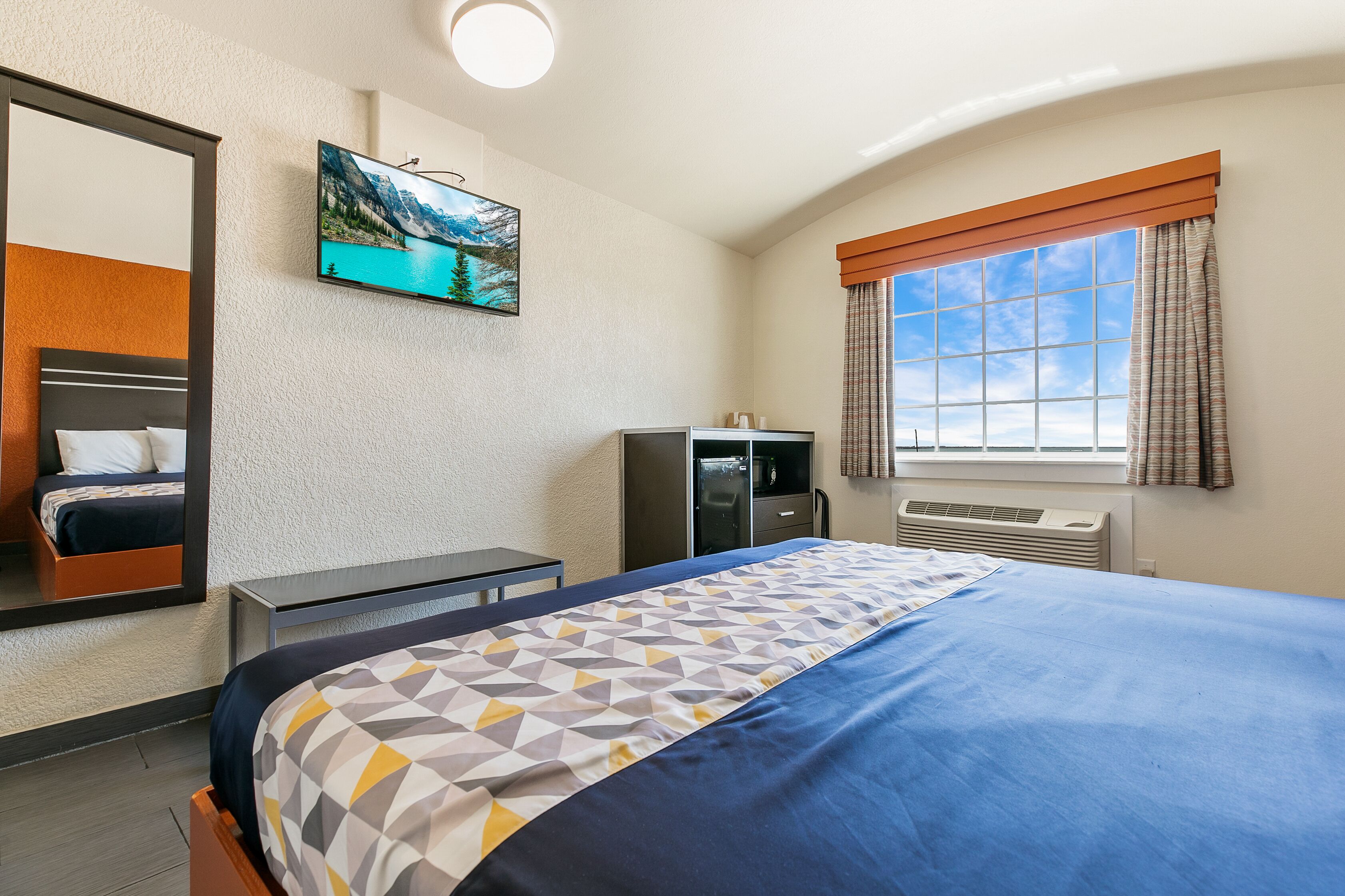 A hotel room with a bed, a desk, a TV mounted on the wall, and a window with curtains.