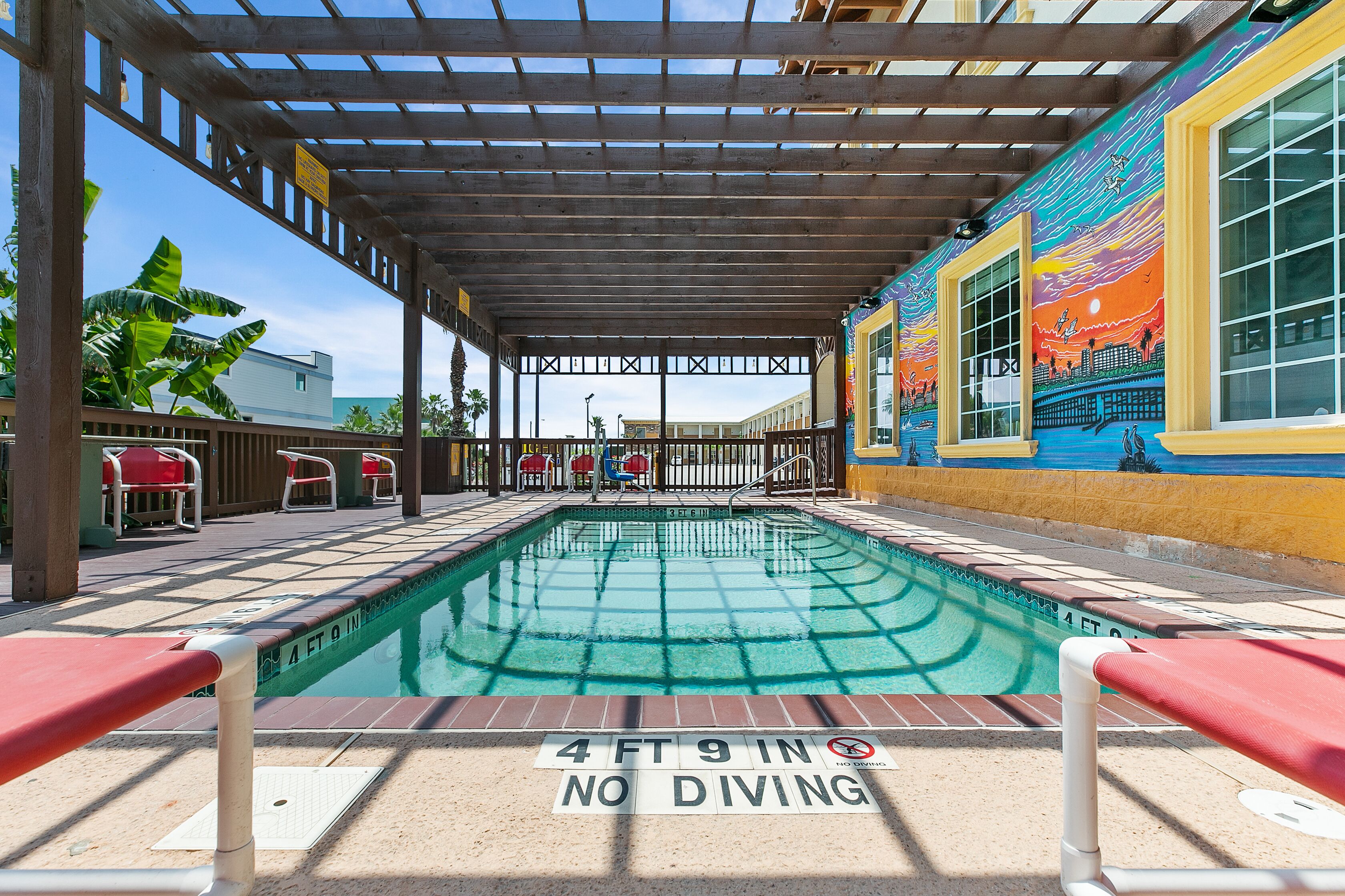 A covered pool area with a '4 FT 9 IN NO DIVING' sign, lounge chairs, and a mural on the wall.
