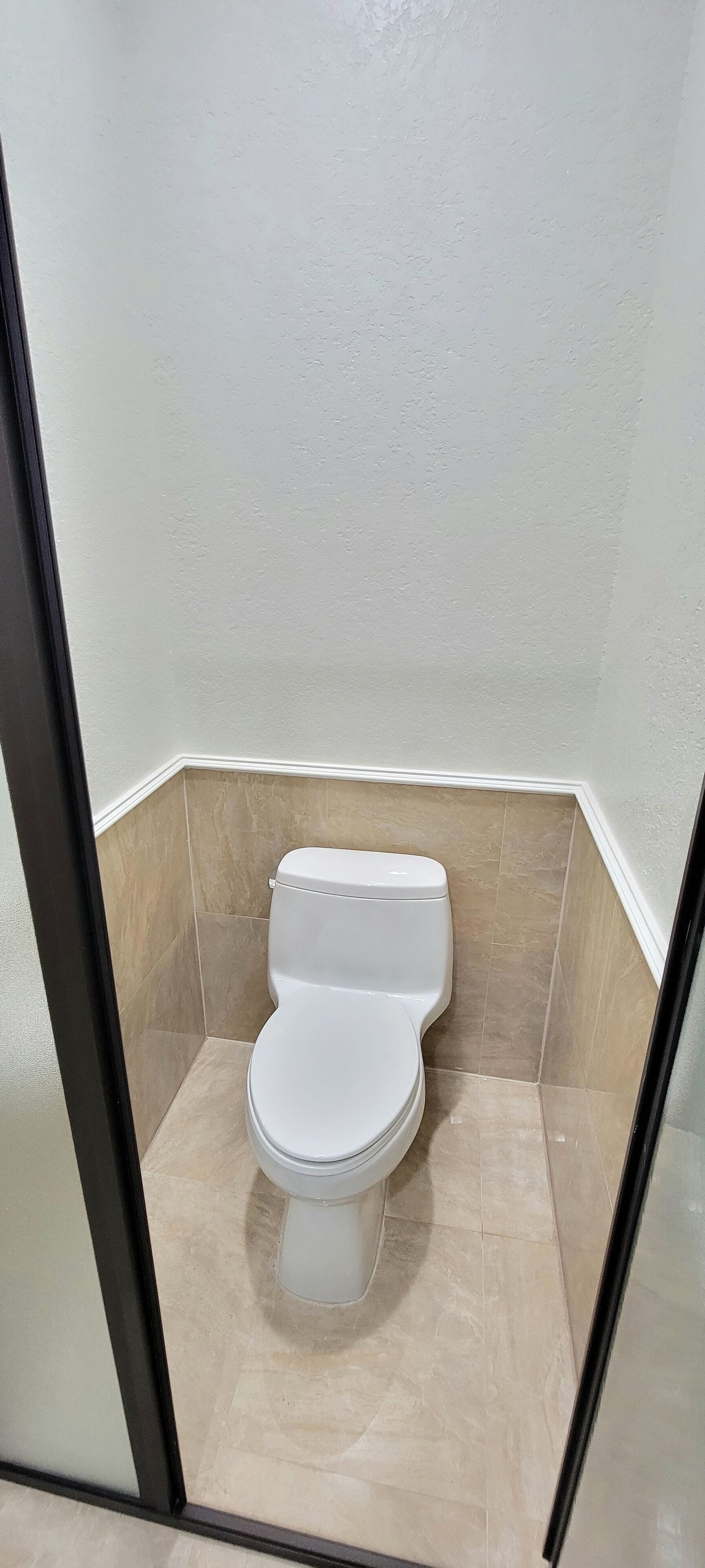 A small, enclosed bathroom with a white toilet, beige tiles on the walls and floor, and a black-framed glass door.
