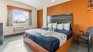 A hotel room with a large bed, a bedside table, a chair, a window with curtains, and an orange accent wall. - Lighthouse Inn (South Padre Island)