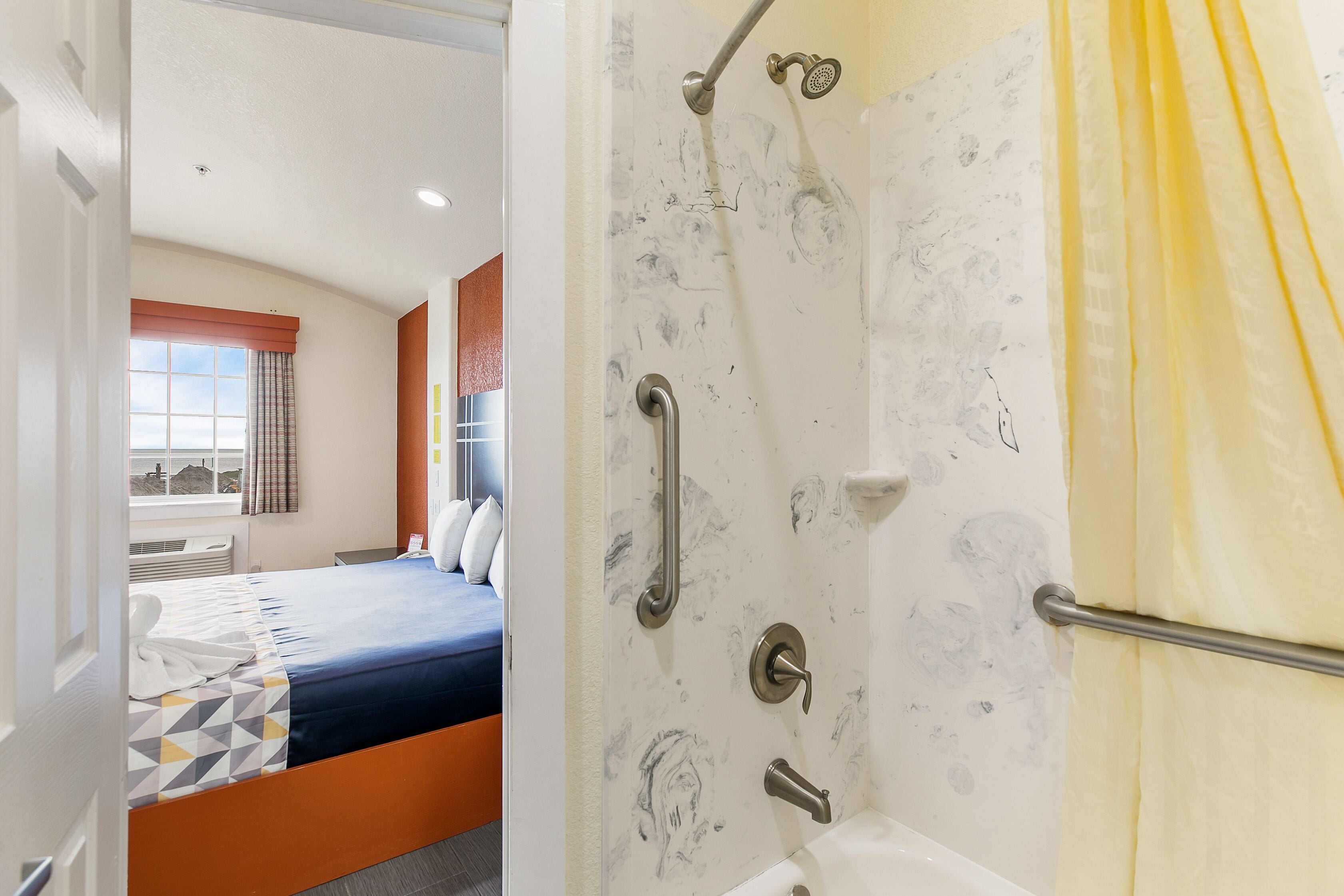 Habitación de hotel con cama, ducha con barras de apoyo y ventana con cortinas.