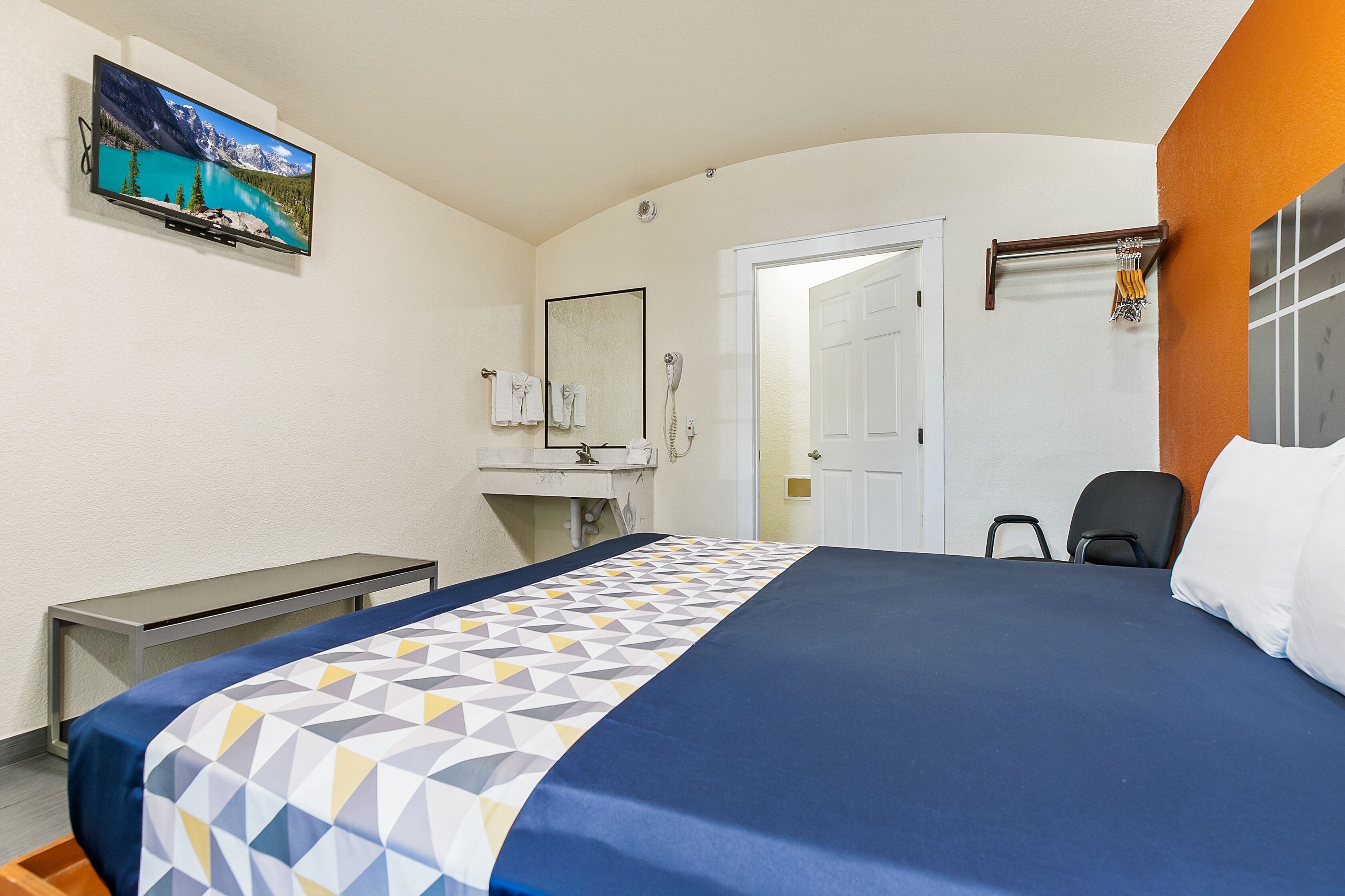 Une chambre d’hôtel comprenant un lit, un bureau, une chaise, une salle de bain avec un lavabo et un miroir, et un téléviseur fixé au mur.