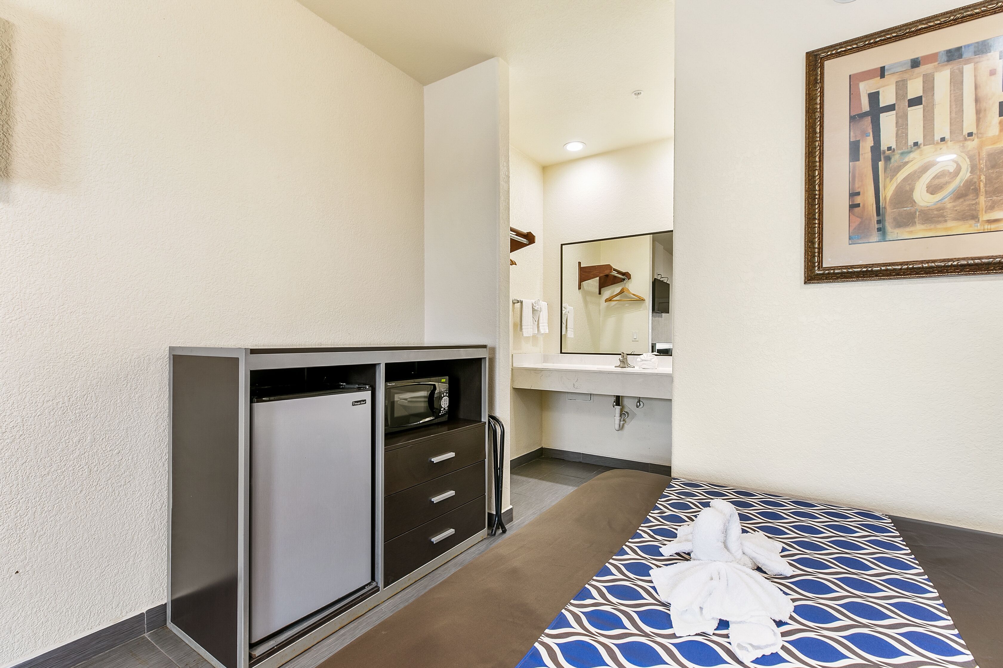A hotel room with a kitchenette, a bed with a blue patterned bedspread, a bathroom with a mirror and towel rack, and a framed abstract painting on the wall.
