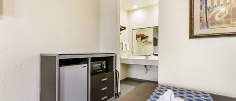 A hotel room with a kitchenette, a bed with a blue patterned bedspread, a bathroom with a mirror and towel rack, and a framed abstract painting on the wall.