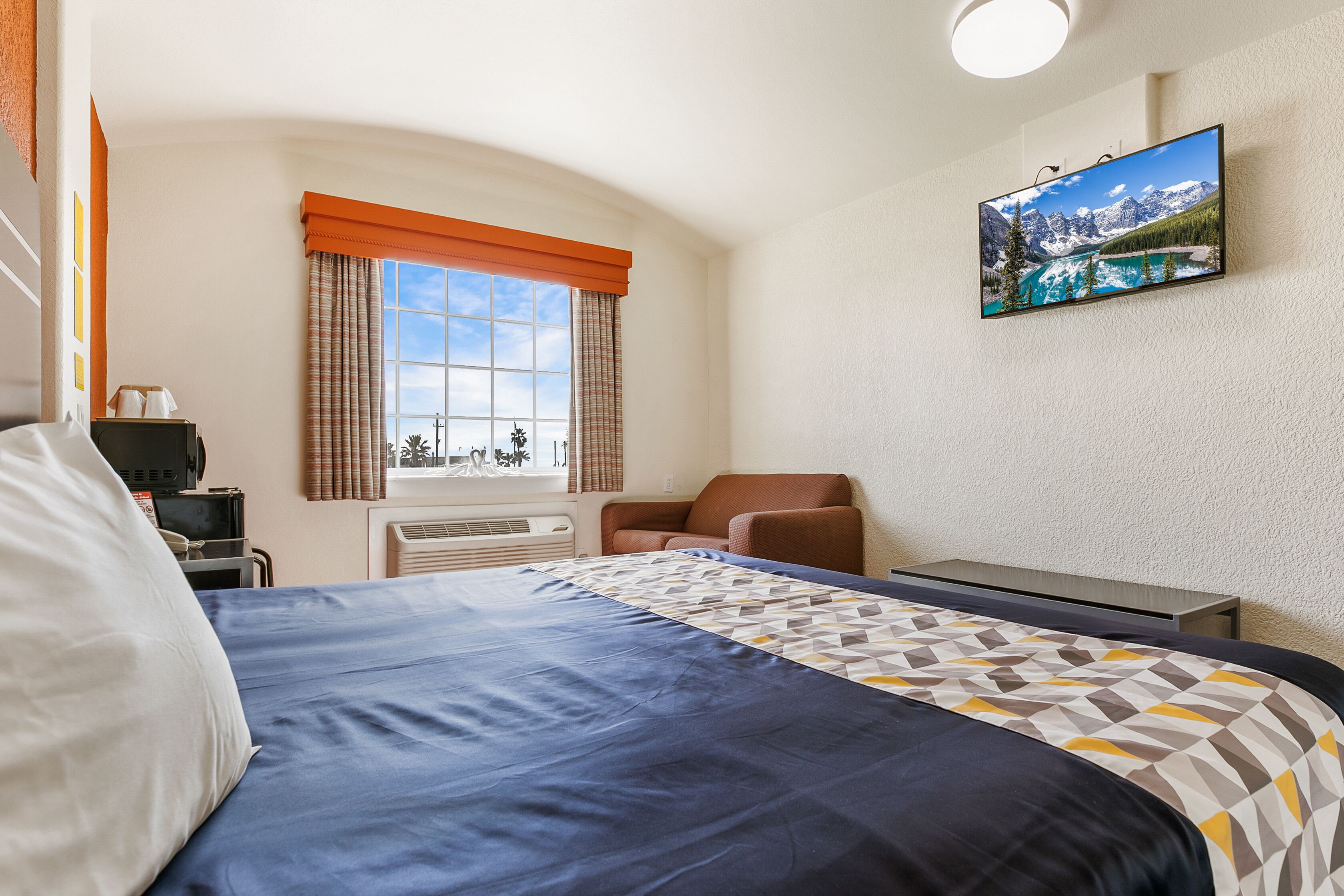 Une chambre d’hôtel avec un lit, un téléviseur fixé au mur, une fenêtre avec des rideaux, une chaise et un bureau avec un ordinateur.