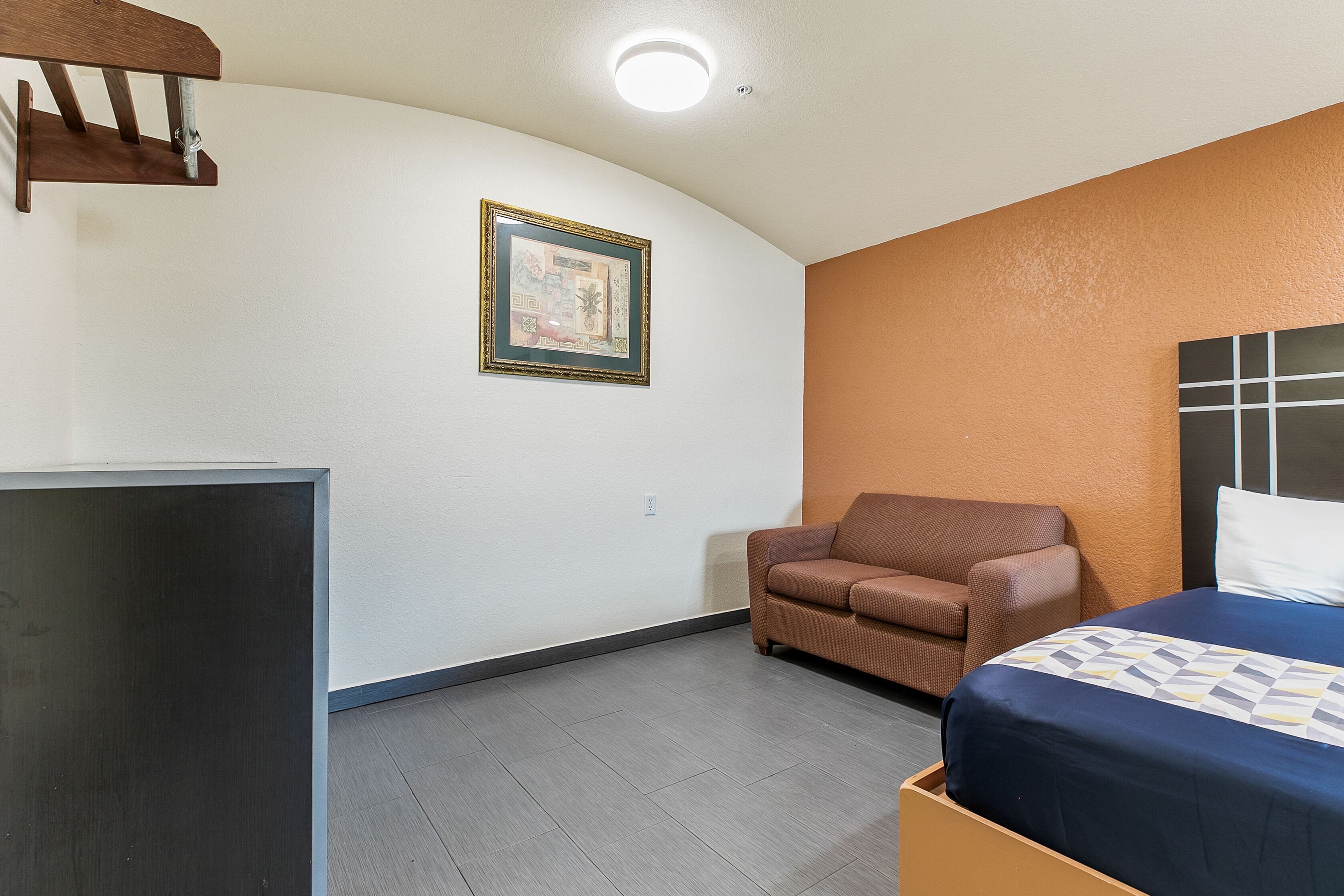 A room with a bed, a chair, a desk, and a framed picture on the wall.