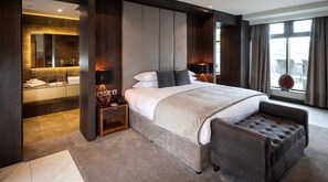 A modern hotel room with a large bed, a seating area, a vanity with a mirror, and a view of the city.