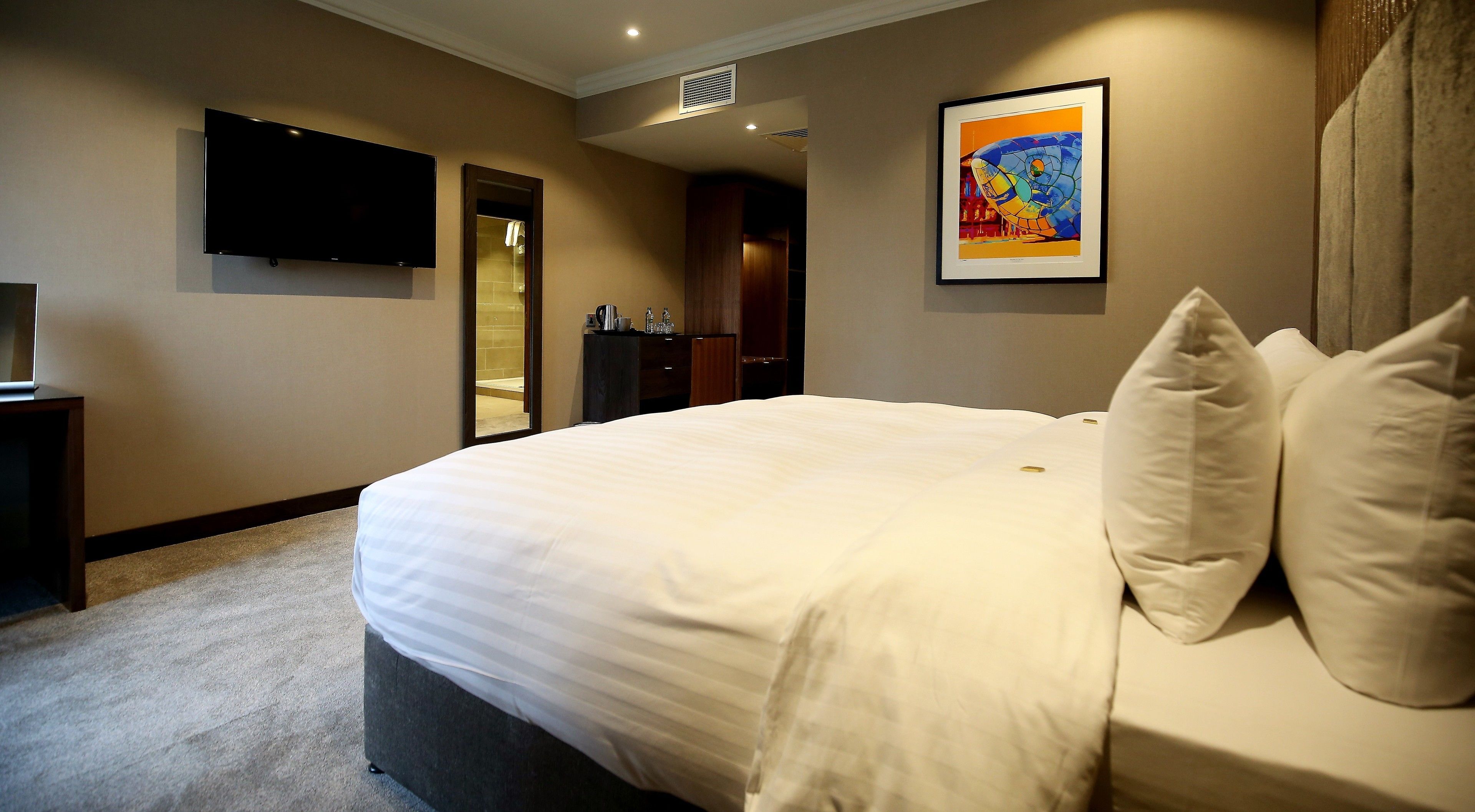 A hotel room with a large bed, a flat-screen TV mounted on the wall, a framed artwork above the bed, and a bathroom visible through an open door.