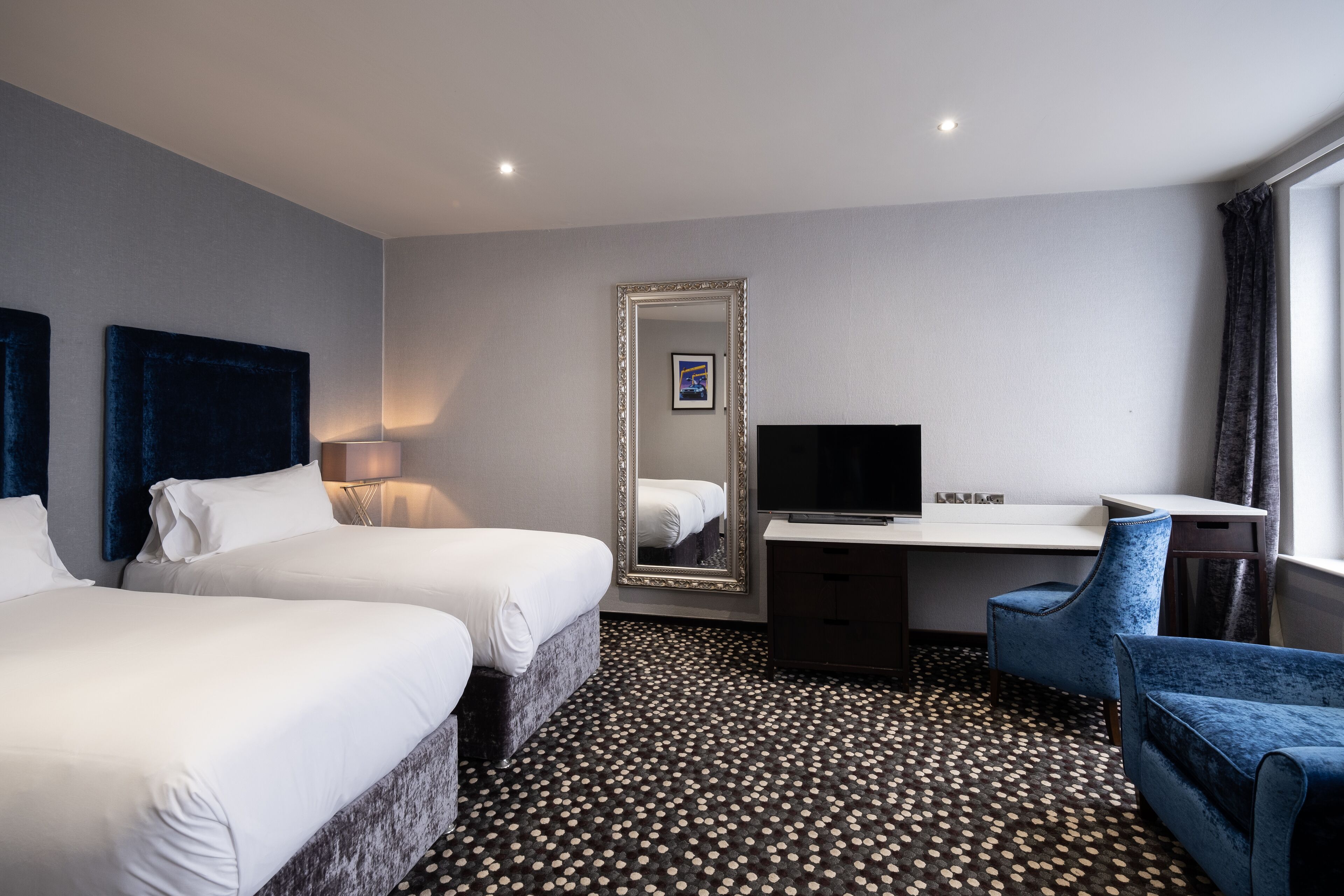 A hotel room with two beds, a desk, a chair, a TV, and a large mirror.
