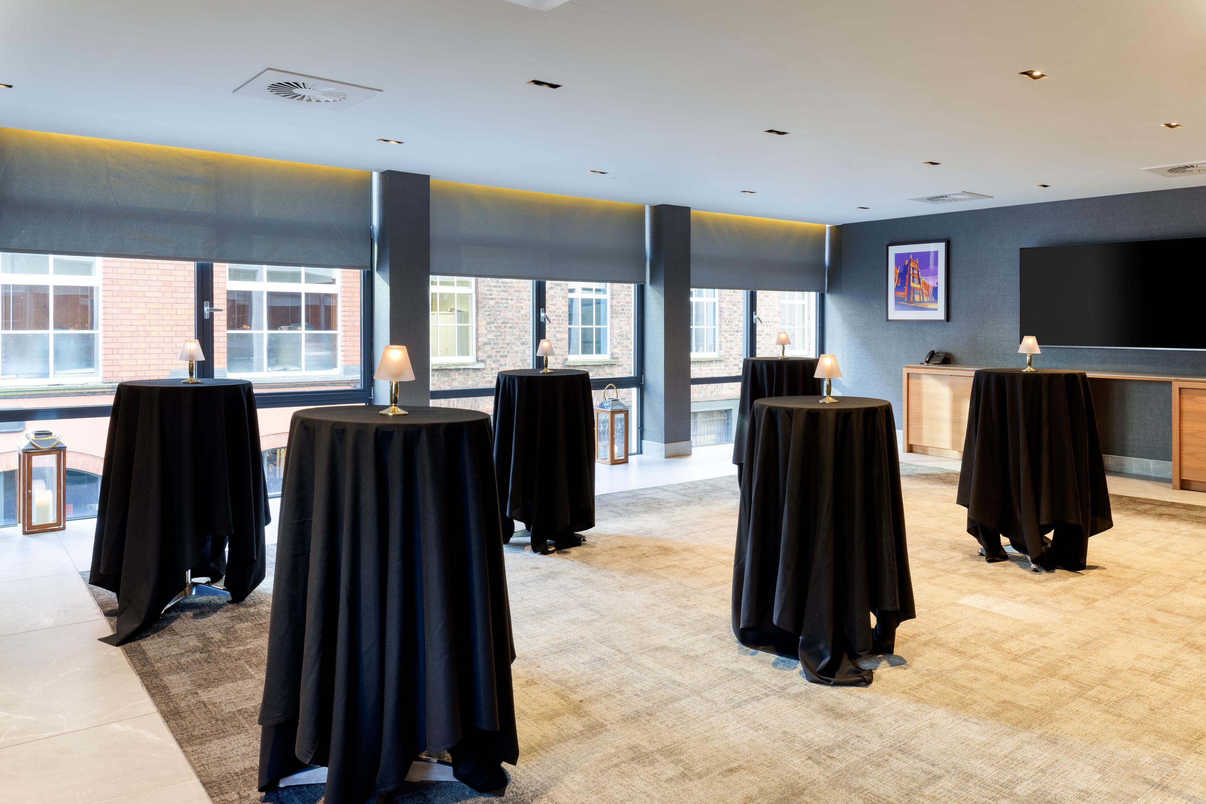 Une chambre d’hôtel avec plusieurs tables rondes recouvertes de nappes noires, une grande fenêtre donnant sur un bâtiment en briques et un téléviseur à écran plat fixé au mur.
