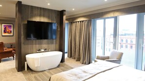Habitación de hotel moderna con una cama grande, una bañera, un televisor de pantalla plana y vistas a la ciudad a través de amplios ventanales.