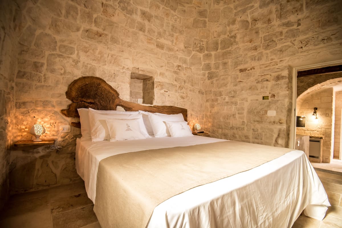 A stone-walled bedroom with a large bed, a wooden headboard, and a stone fireplace.