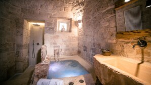 A stone-walled bathroom with a stone bathtub, a wooden shelf, and a small window. - La Corte dell'Astore (Alberobello)