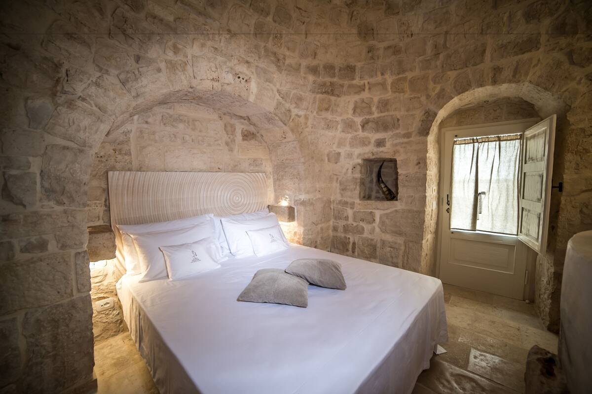 A stone-walled bedroom with a bed, pillows, a nightstand, and a window with curtains.