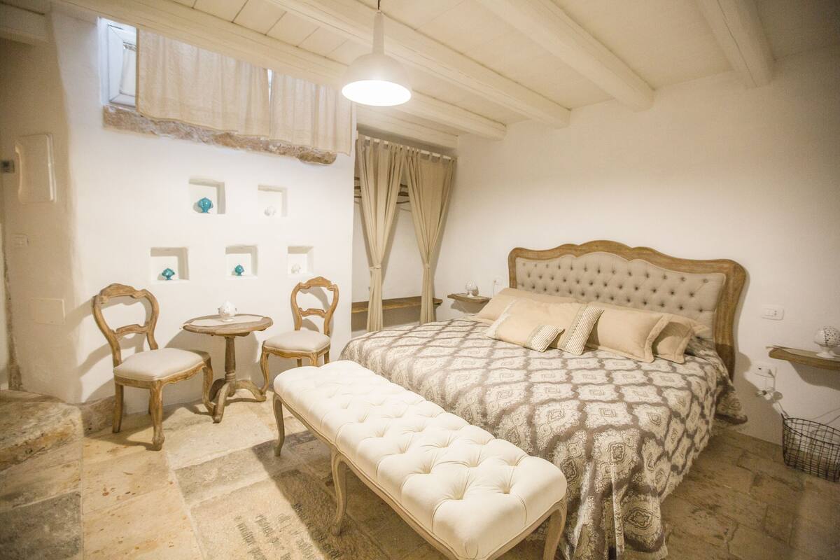 A bedroom with a large bed, a bench, two chairs, a small table, and a window with curtains.