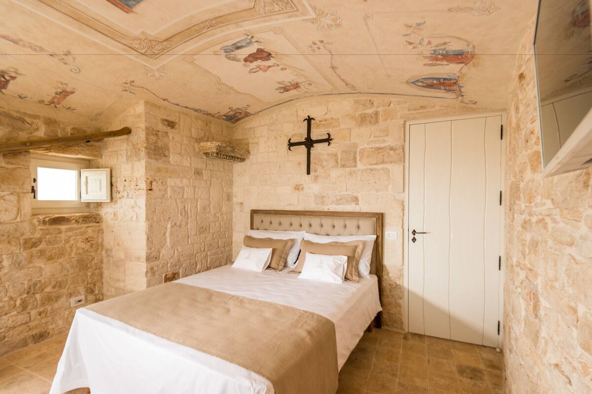 A bedroom with a stone wall, a bed with beige bedding, a wooden headboard, and a cross on the wall.