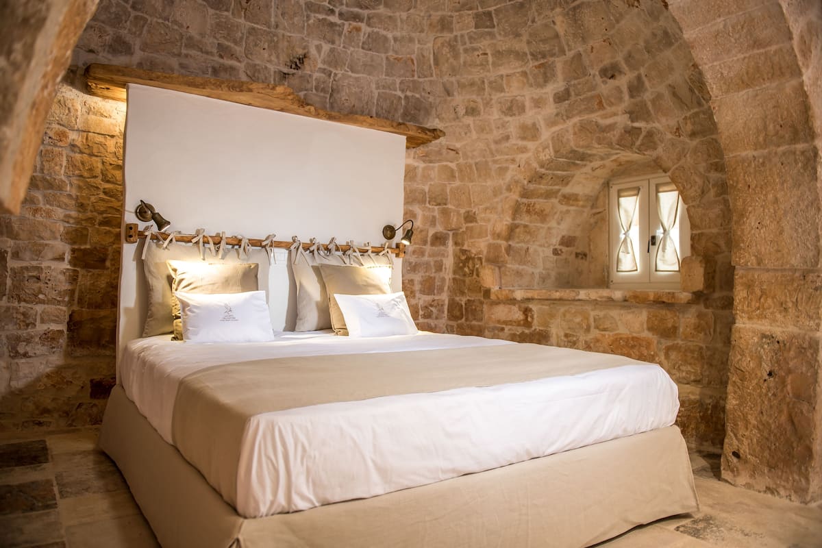 A stone-walled bedroom with a large bed, wooden headboard, and a small window with curtains.