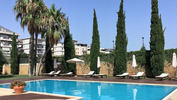 Piscina com espreguiçadeiras e guarda-sóis, rodeada por ciprestes altos e um edifício ao fundo.