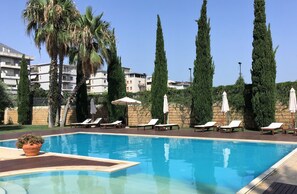 Une piscine entourée de chaises longues et de parasols, bordée de grands cyprès et avec un bâtiment visible au loin.