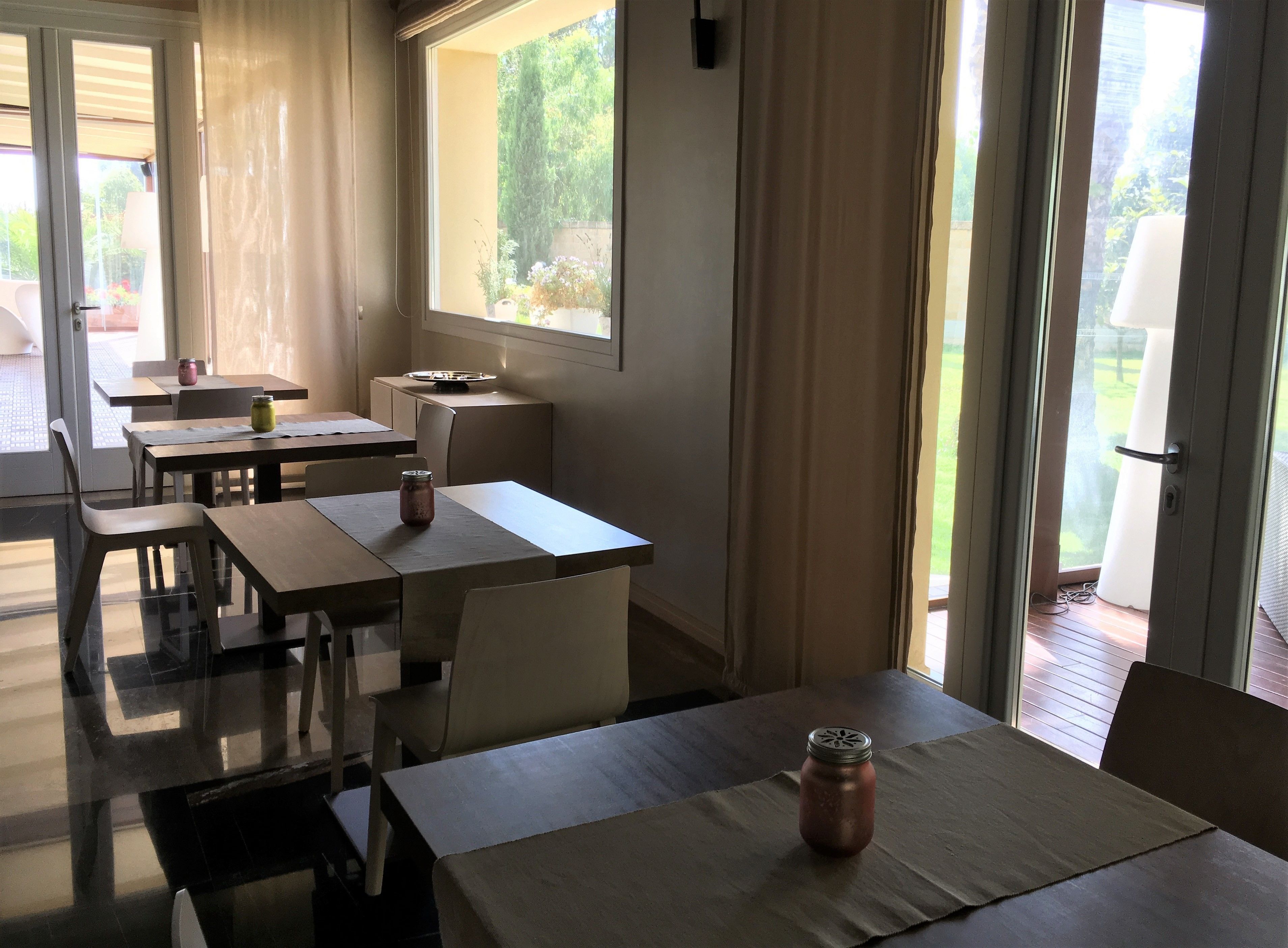 A dining area with tables and chairs, a window with curtains, and a view of greenery outside.
