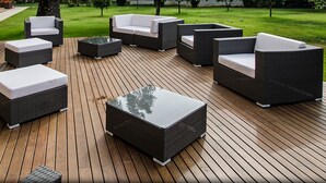 Um pátio moderno ao ar livre com móveis de vime e uma mesa com tampo de vidro, rodeado por um deck de madeira e um gramado bem cuidado.