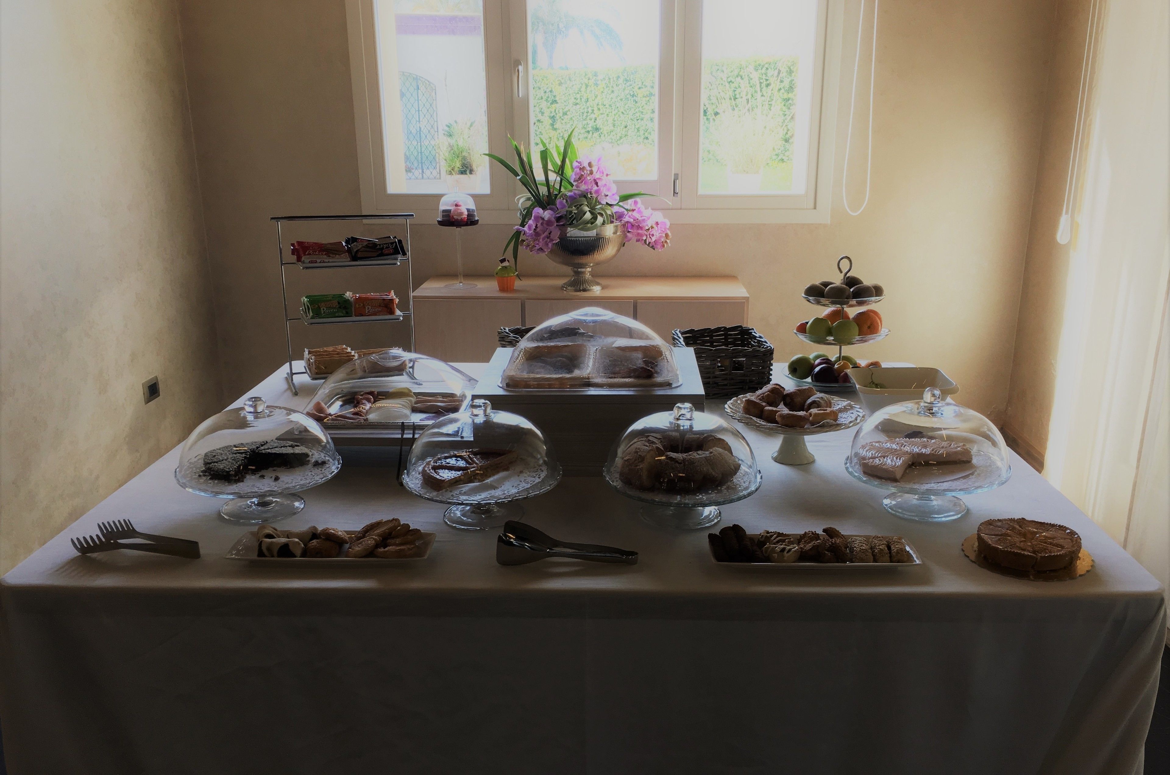 A table with various desserts and pastries, a small shelf with snacks, and a window with a view of greenery.