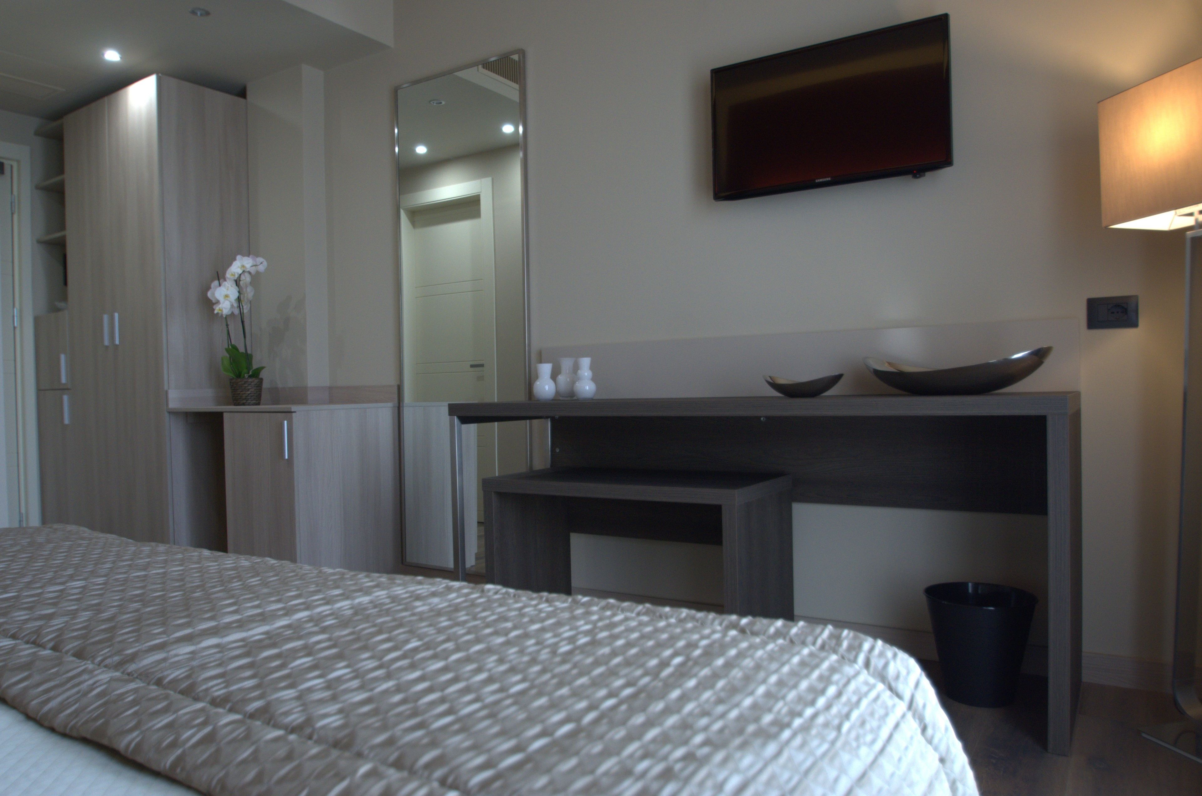 A bedroom with a bed, a bedside table, a mirror, a TV, and a small desk with a bowl and a trash can.