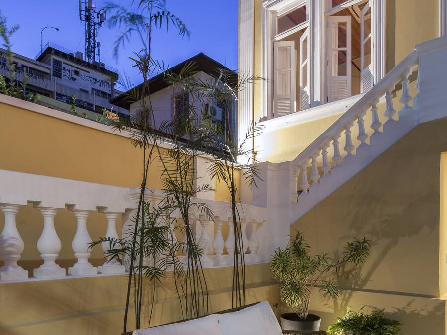 Un patio con un sofá blanco, mobiliario de mimbre, plantas en macetas y una escalera que lleva a un nivel superior.
