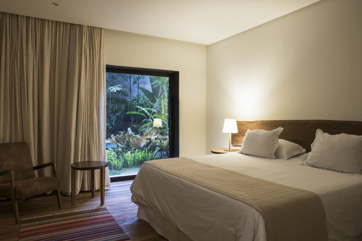 A hotel room with a large bed, a chair, a small table, and a view of greenery through a window.
