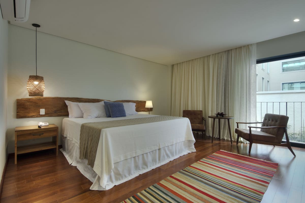 A hotel room with a large bed, a wooden nightstand, a small table, a chair, and a view of the outside through a window.