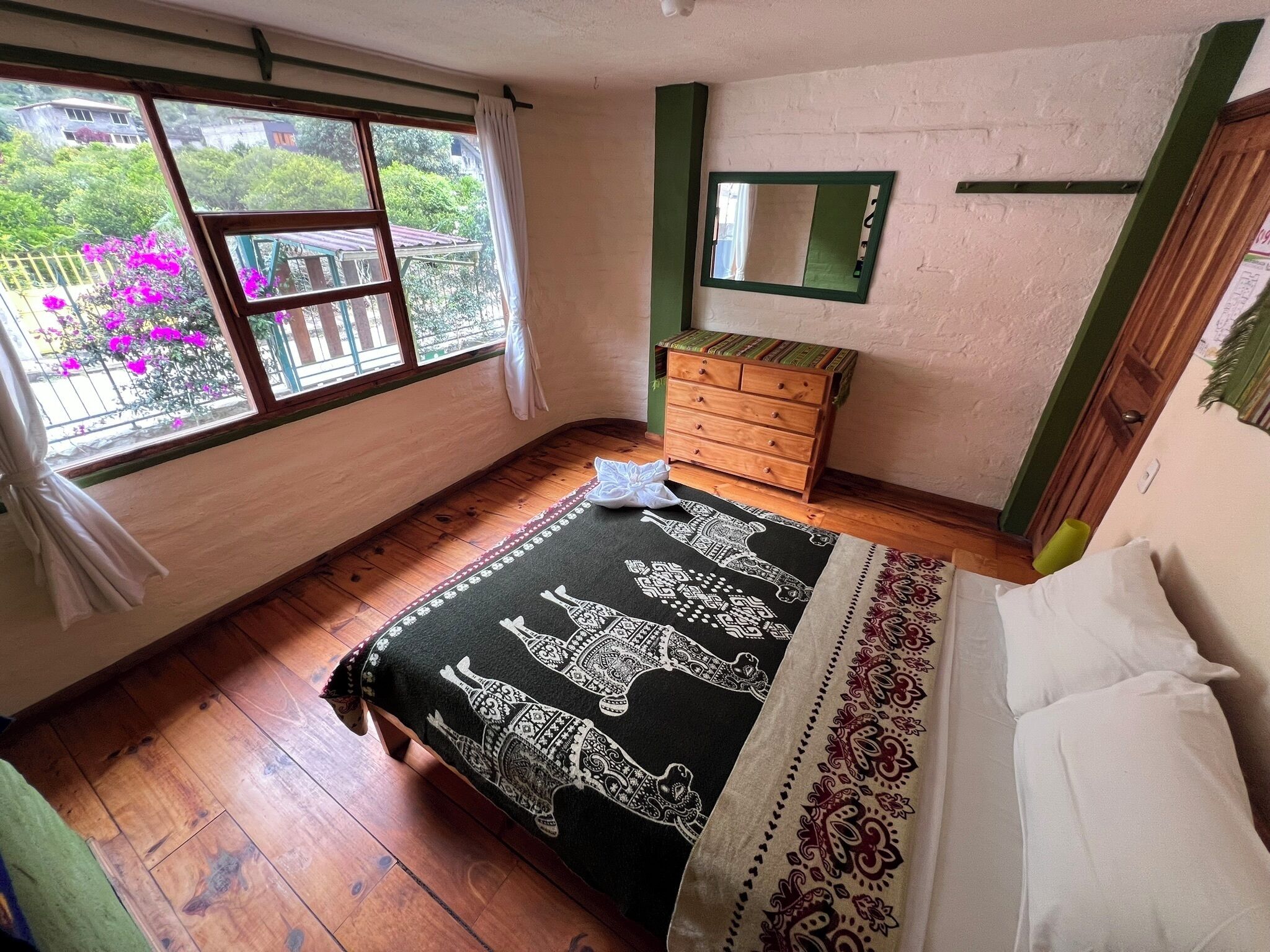 Un dormitorio con piso de madera, una cama con ropa de cama blanca, un aparador de madera, una ventana con cortinas y un espejo en la pared.