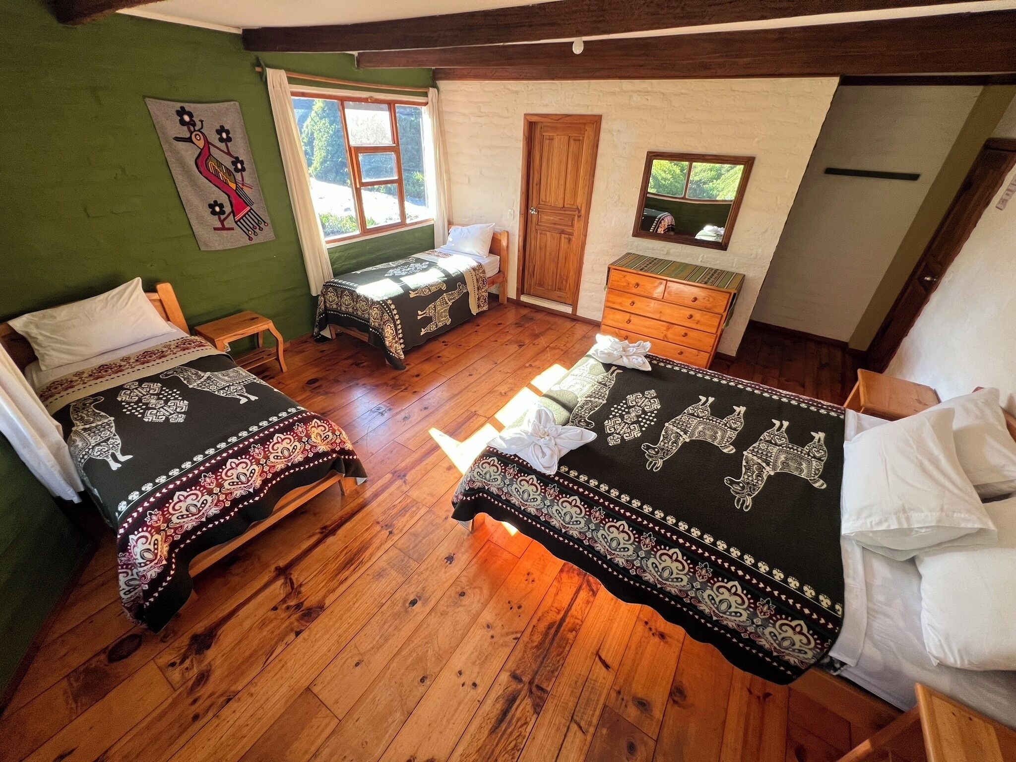 A room with two beds, wooden flooring, a wooden dresser, a window with curtains, and a green wall with a wall hanging.