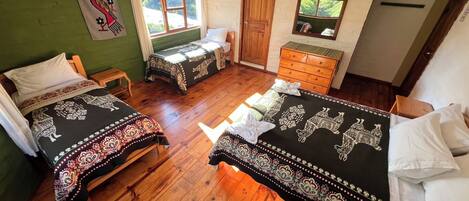 A room with two beds, wooden flooring, a wooden dresser, a window with curtains, and a green wall with a wall hanging.