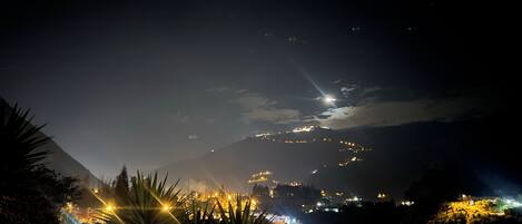 Vista nocturna de una zona montañosa con edificios iluminados y una brillante luna en el cielo.