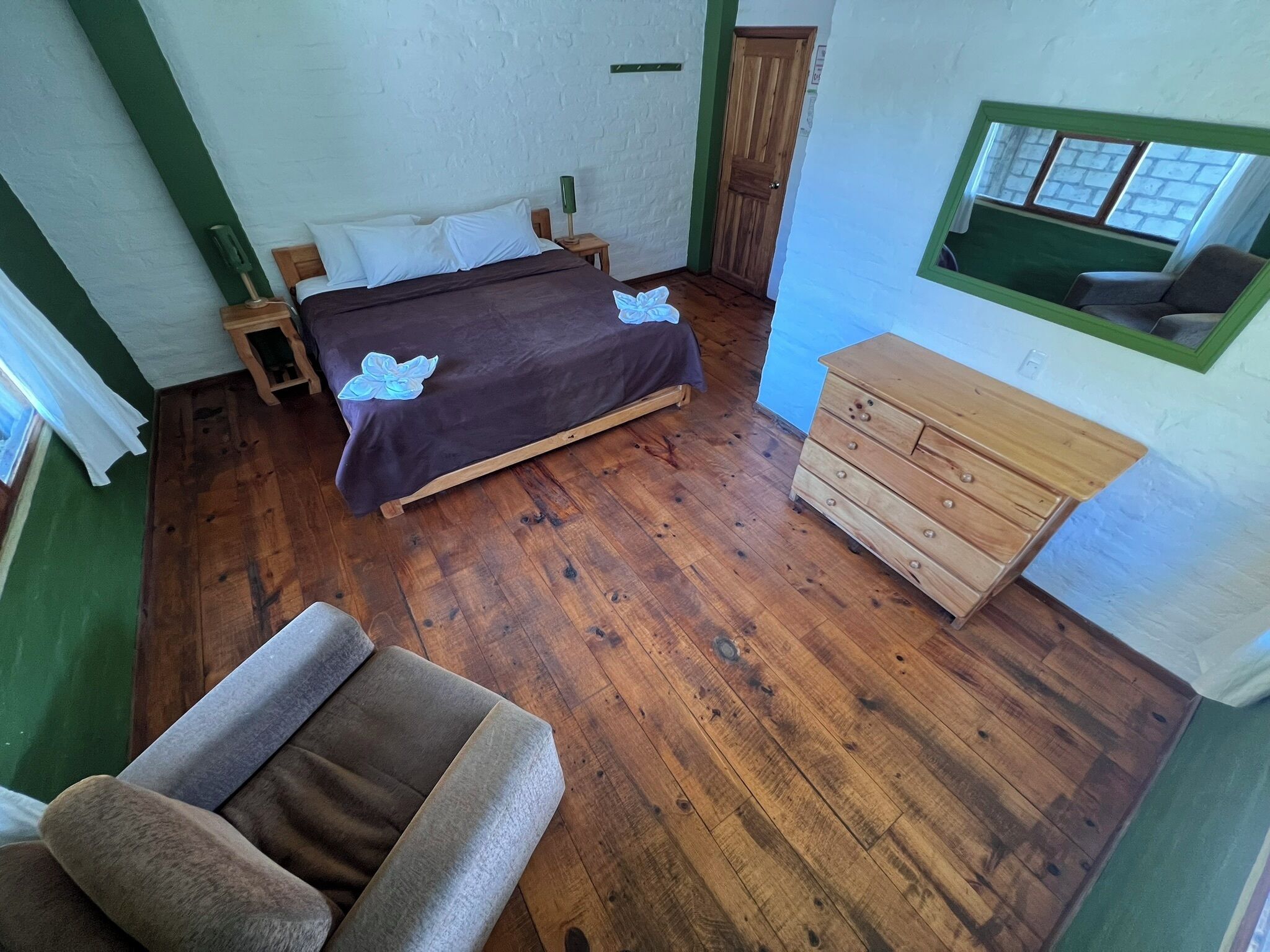 A bedroom with a wooden floor, a bed with white linens, a wooden dresser, and a window with a view.