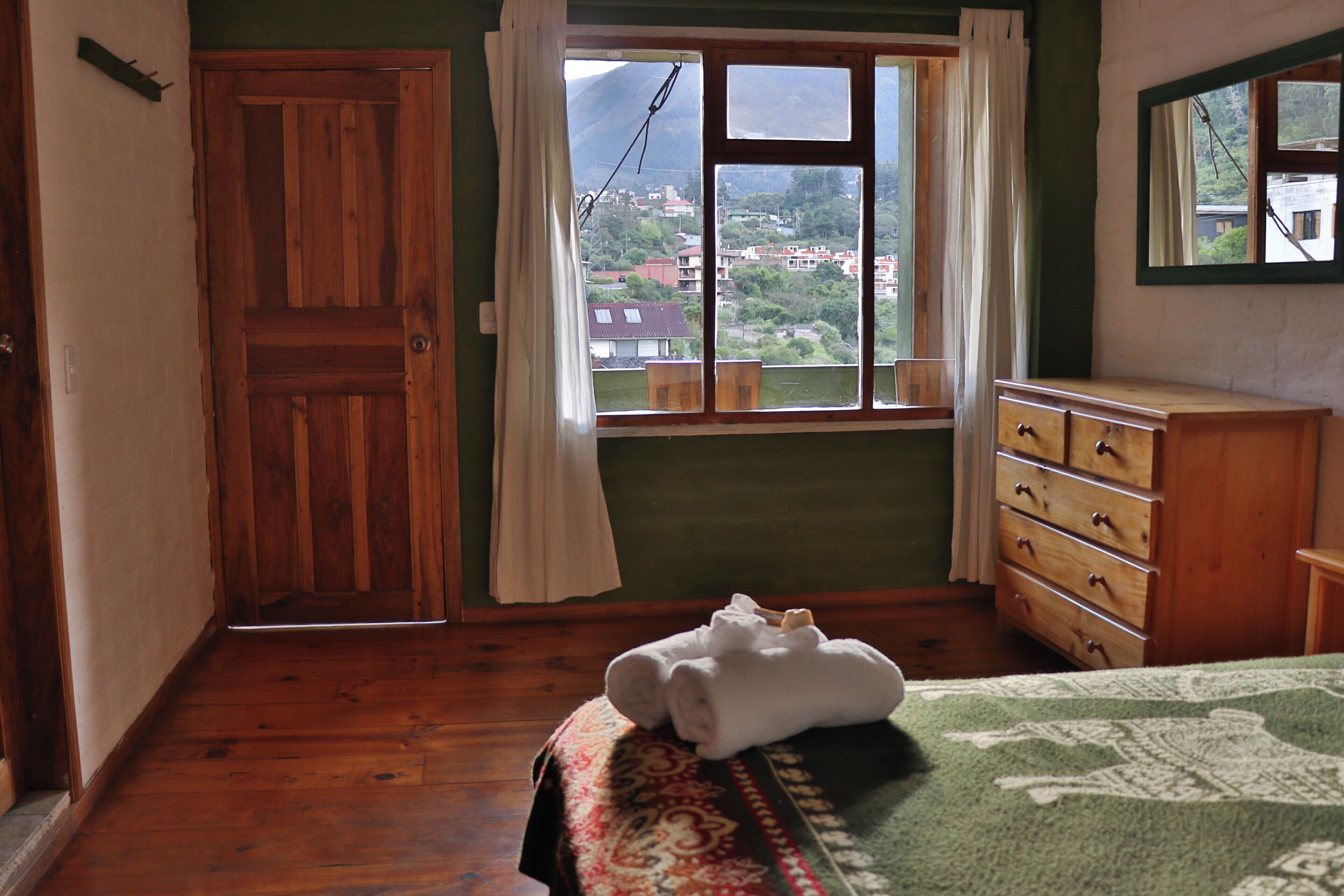 A room with a wooden door, a window with curtains, a dresser, and a bed with a patterned bedspread.
