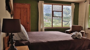 Een slaapkamer met een bed, nachtkastje, stoel en een raam met uitzicht op een bergachtig landschap.