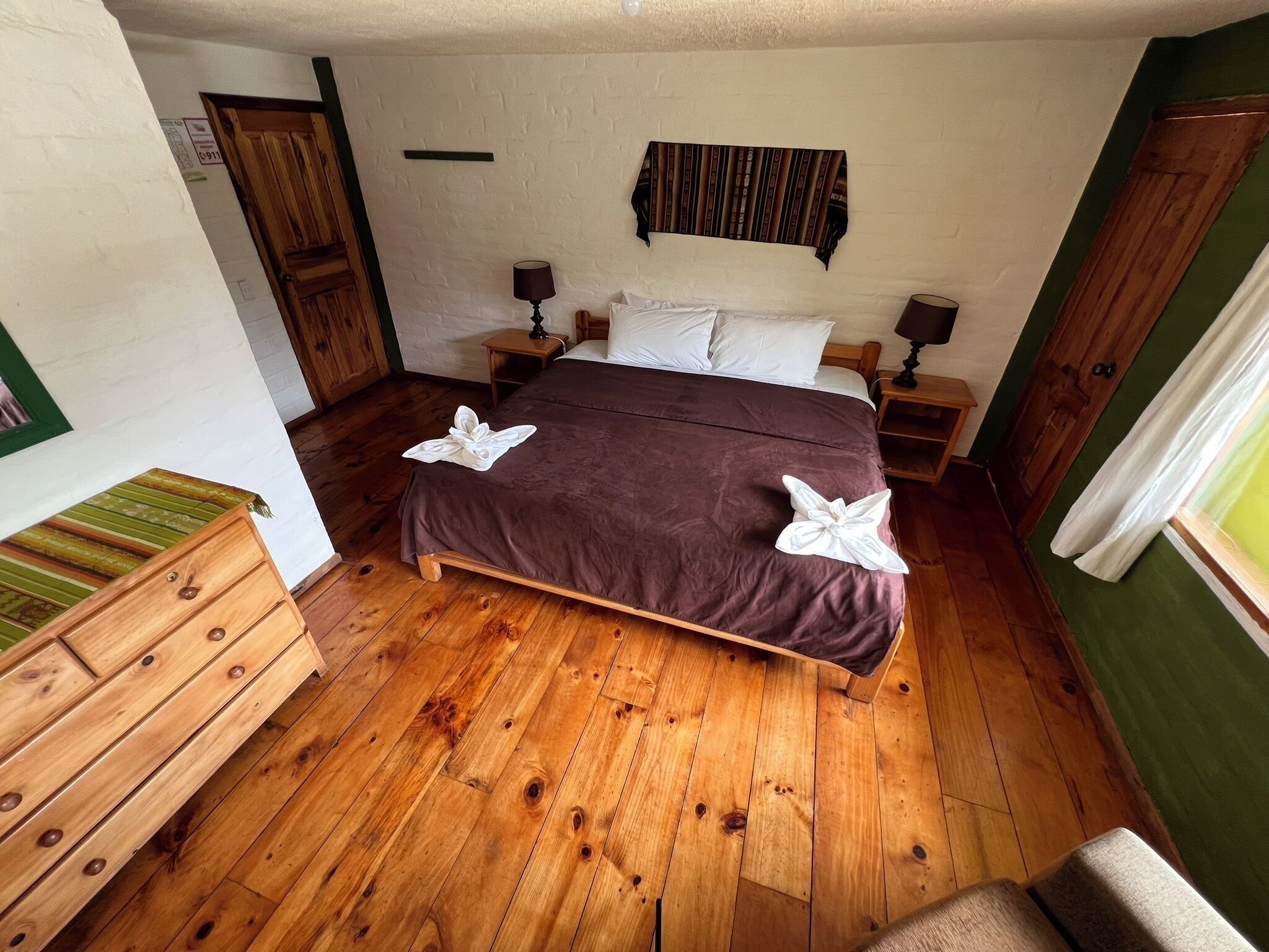 Una camera da letto con pavimento in legno, un letto con copriletto marrone, due comodini con lampade, una cassettiera in legno e una parete verde.