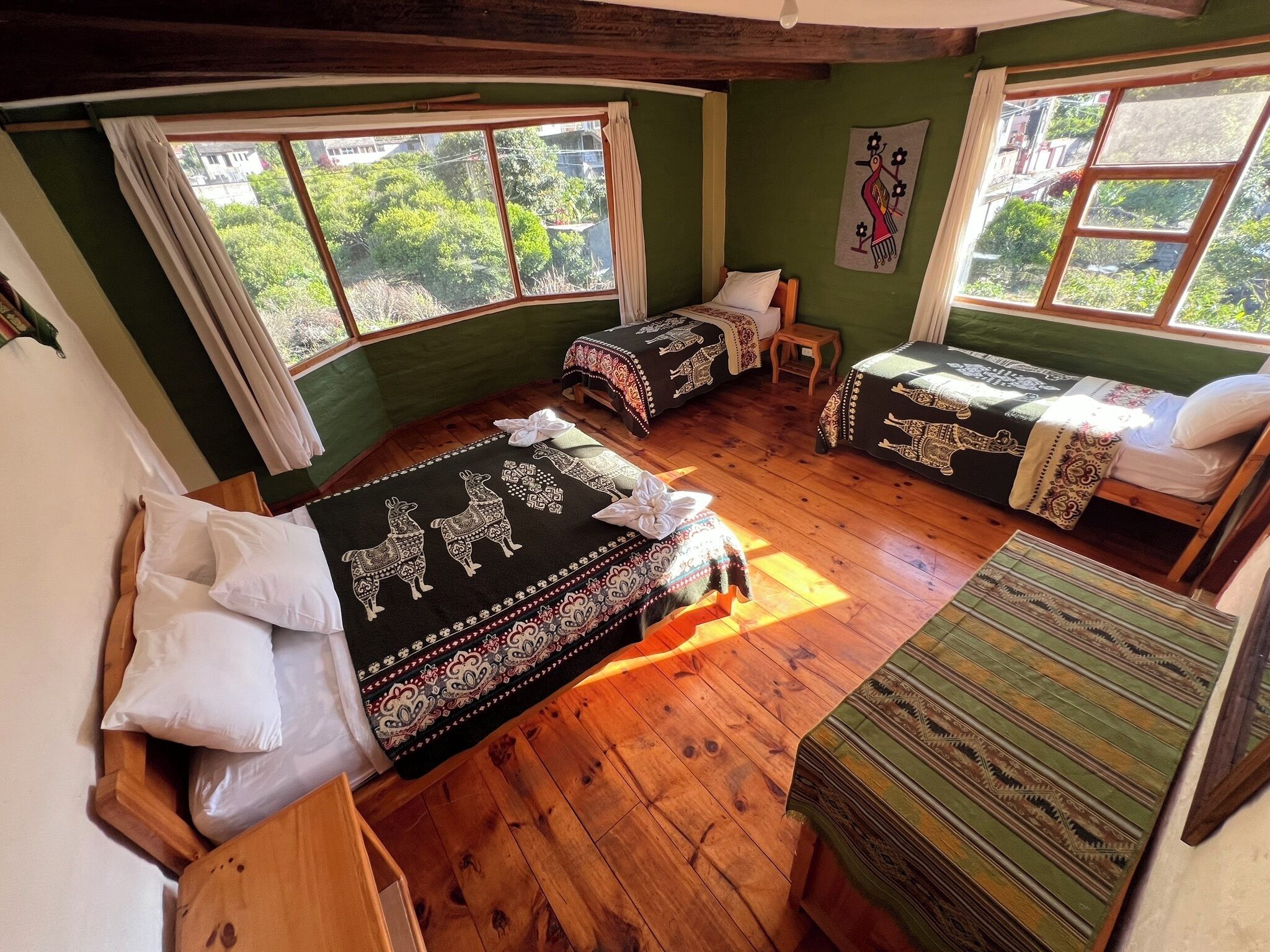 A room with two beds, a wooden floor, and large windows with a view of greenery.