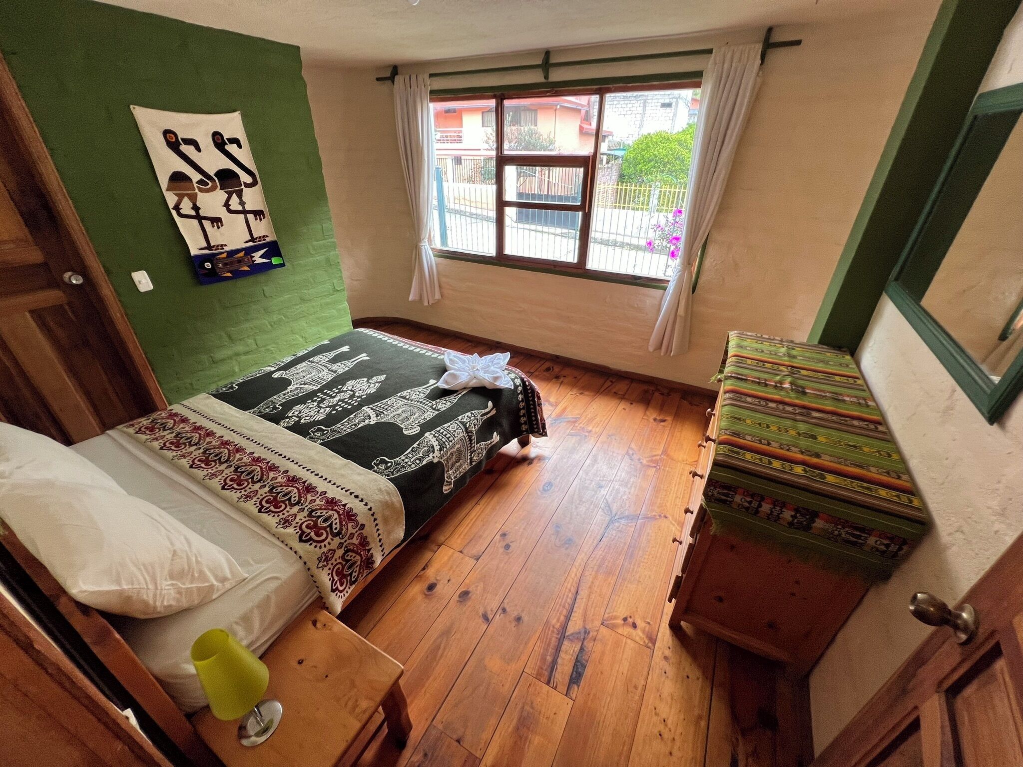 A small, cozy room with a bed, a wooden floor, a window with curtains, and a green wall.