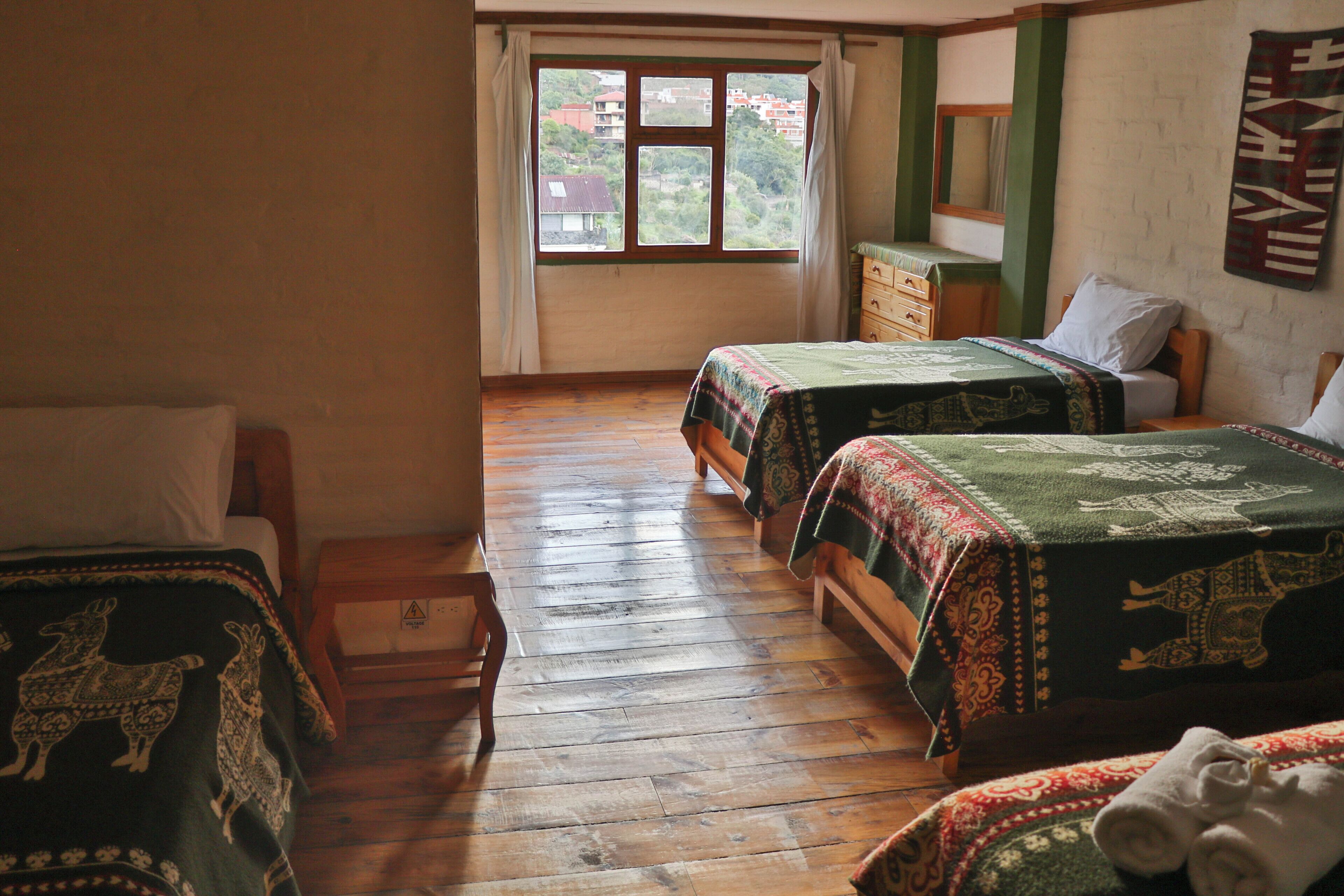 A room with two beds, wooden flooring, a window with curtains, a dresser, and a wall tapestry.