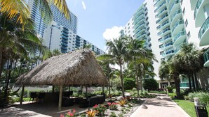 A resort area with a thatched-roof structure, palm trees, and modern high-rise buildings in the background. - The Tides by MiaRentals (Hollywood)