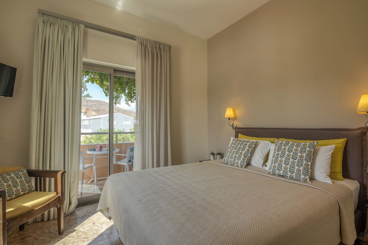 A hotel room with a large bed, a wooden chair, a balcony with a table and chairs, and a TV mounted on the wall.