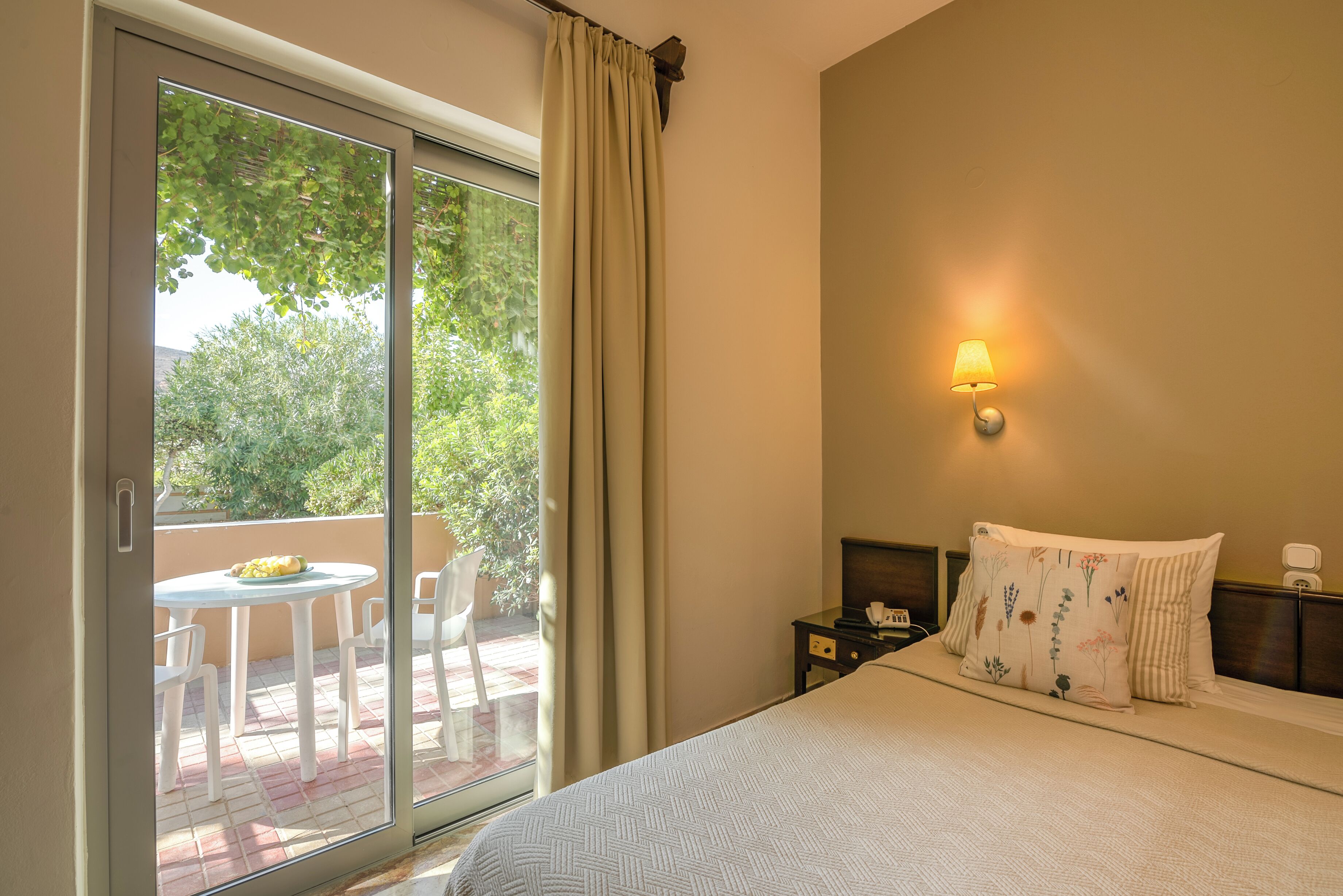 Habitación de hotel con cama, mesita de noche, lámpara, ventana con cortinas y vista a un patio exterior con sillas y mesa.