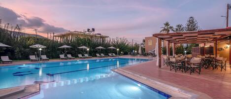 A swimming pool with lounge chairs and umbrellas, surrounded by a covered patio area with tables and chairs.
