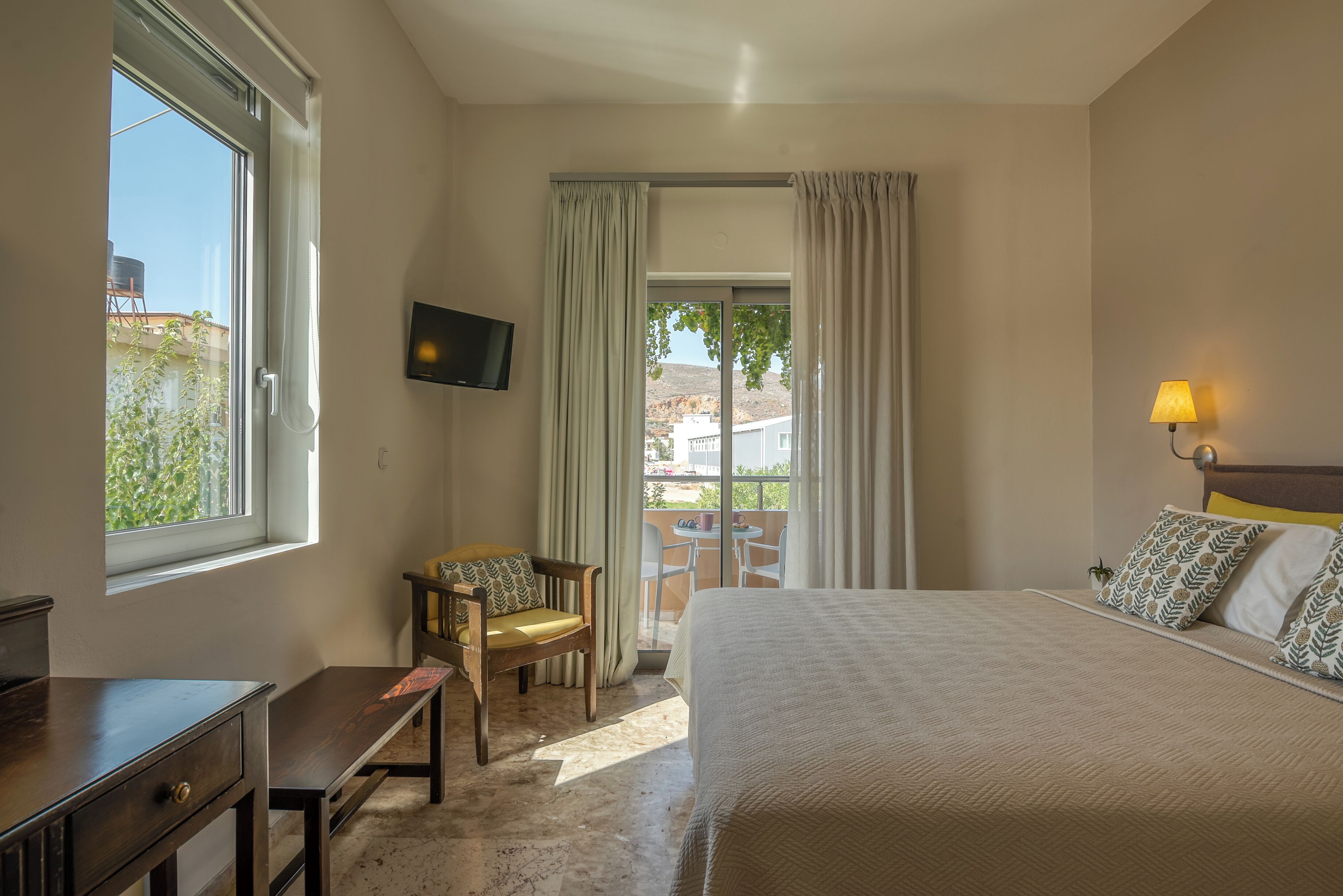 Ein Schlafzimmer mit Bett, Schreibtisch, Sessel, Fernseher und einem Balkon mit Vorhängen.