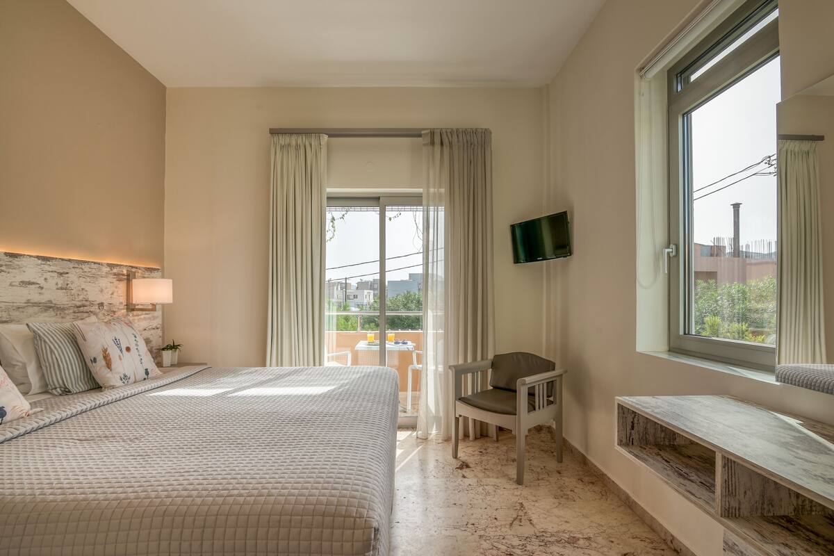 A bedroom with a bed, a chair, a TV, a balcony with curtains, and a view of the outside.
