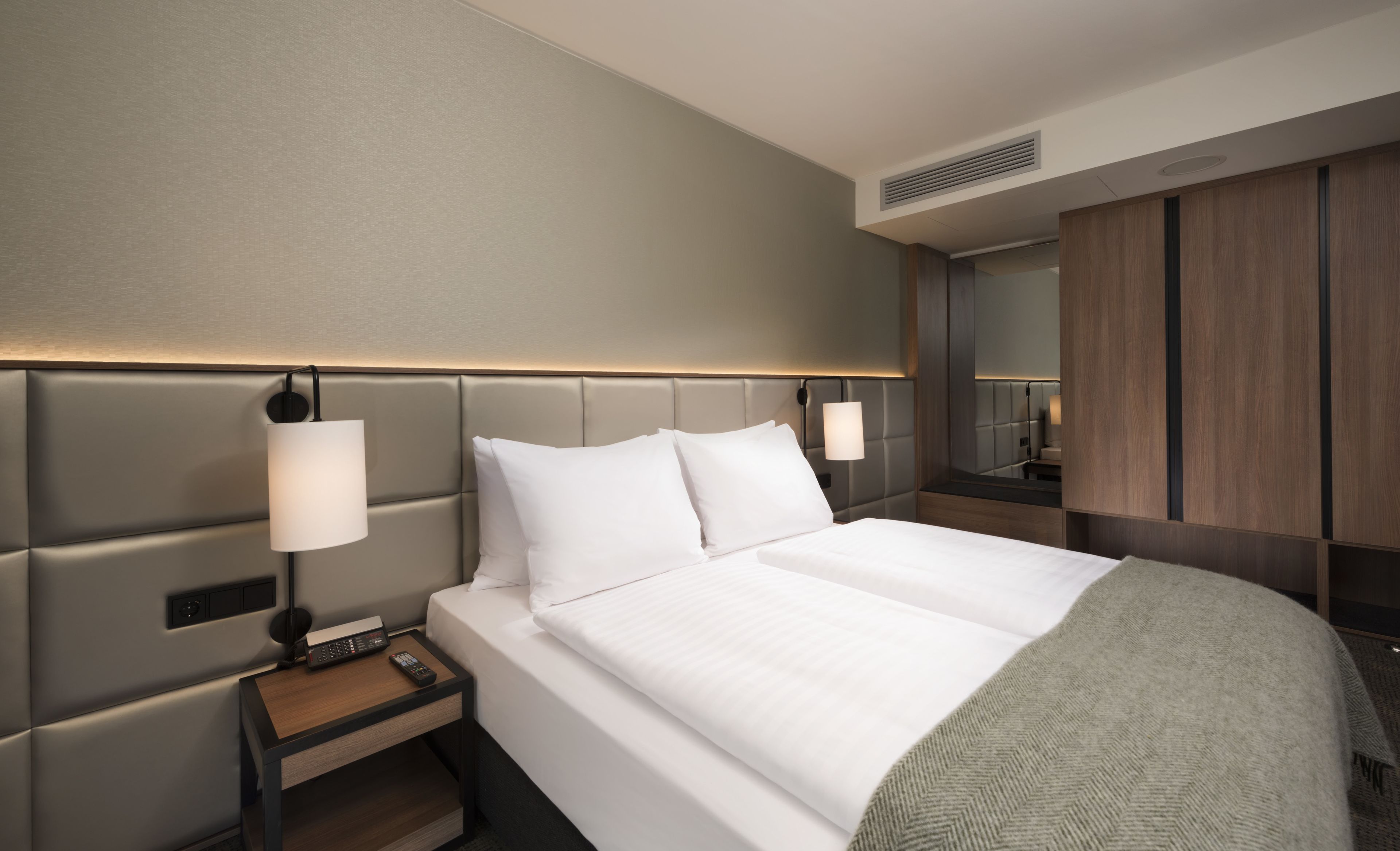 A modern hotel room with a large bed, a nightstand with a lamp, a remote control, and a mirror on the wall.
